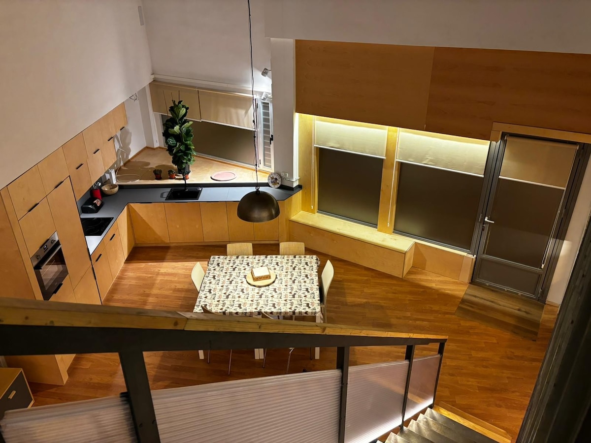 An open kitchen area features light wooden cabinetry and modern appliances. A dining table is surrounded by six chairs, positioned near large windows with soft blinds. The warm wooden flooring adds a cozy touch to the space, illuminated by overhead lighting.