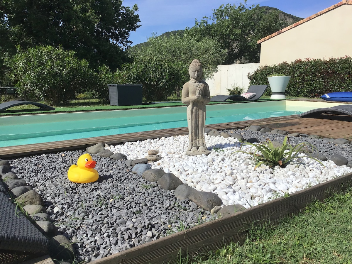 A serene outdoor space features a stone statue standing amidst a decorative garden with white and gray pebbles. A small rubber duck adds a touch of whimsy to the scene, which is bordered by a clear swimming pool and green landscaping.