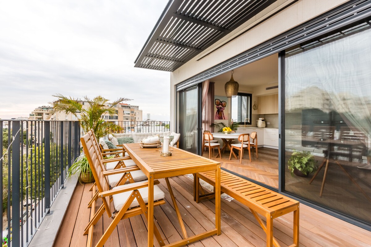 A spacious private terrace is featured, furnished with a large wooden dining table and several matching chairs. Sunlight filters through the modern awning, casting soft shadows across the wooden floor. Lush plants are positioned strategically, adding a touch of greenery to the outdoor space.