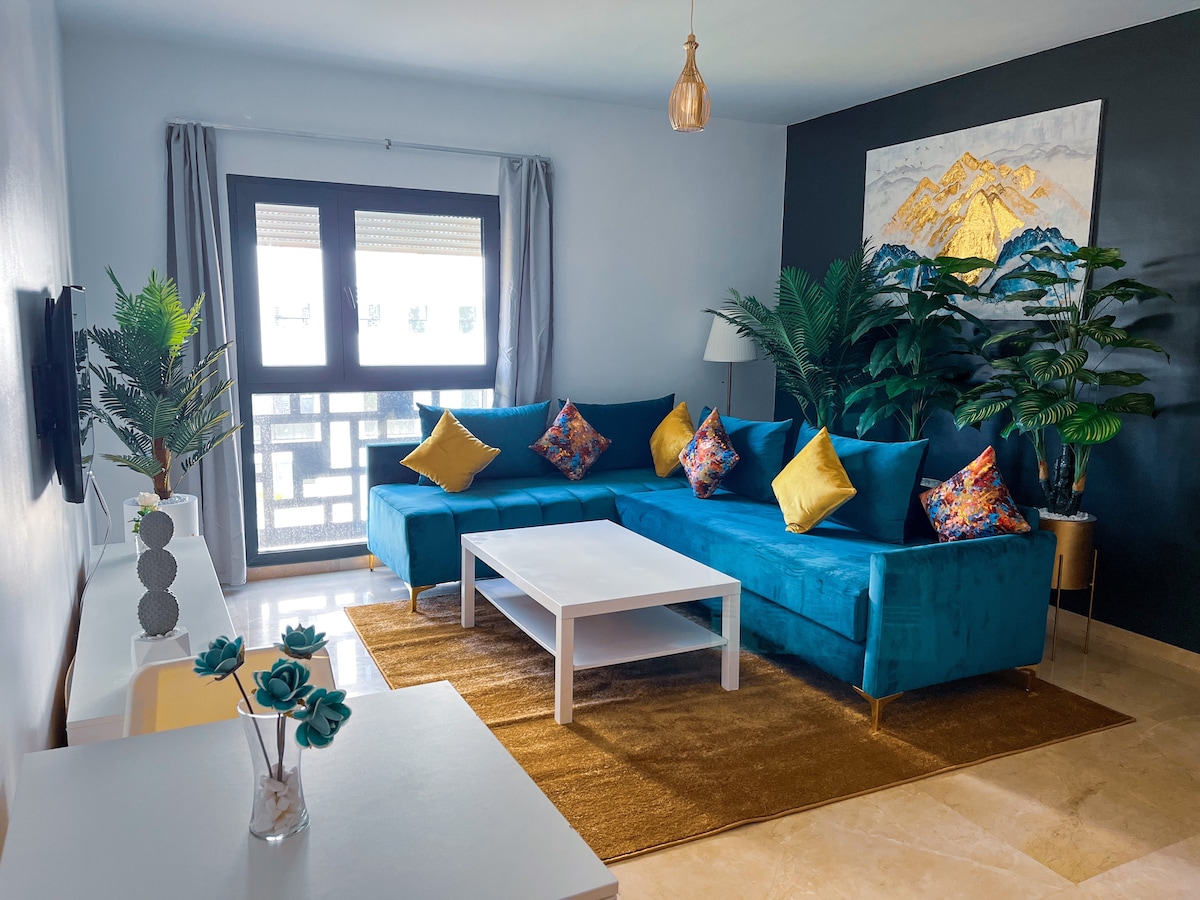 A spacious living area is highlighted by a large turquoise sectional sofa adorned with decorative yellow cushions. A white coffee table is placed on a soft area rug, complemented by a painting of mountains. Natural light filters through large glass doors, enhancing the decor.