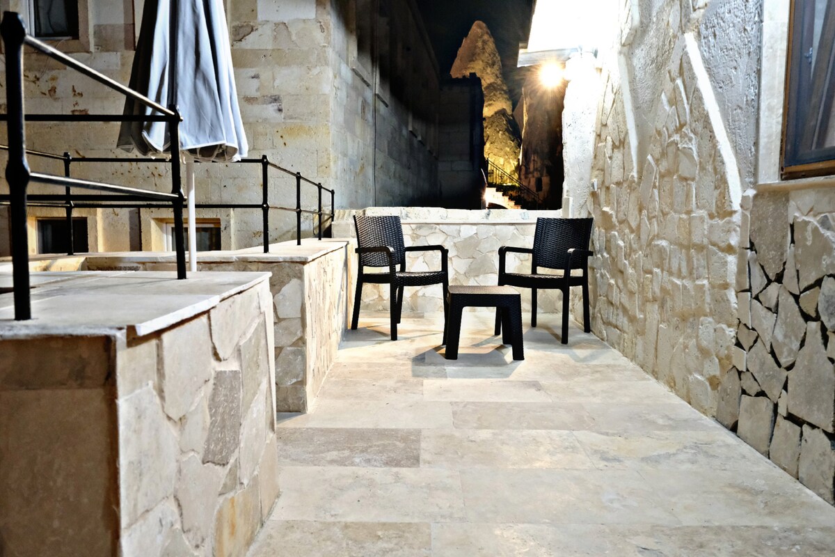 An outdoor seating area features two black chairs positioned on a natural stone patio. A small black table is centered between the chairs. The space is illuminated by soft lighting, creating a cozy ambiance against the backdrop of textured stone walls.