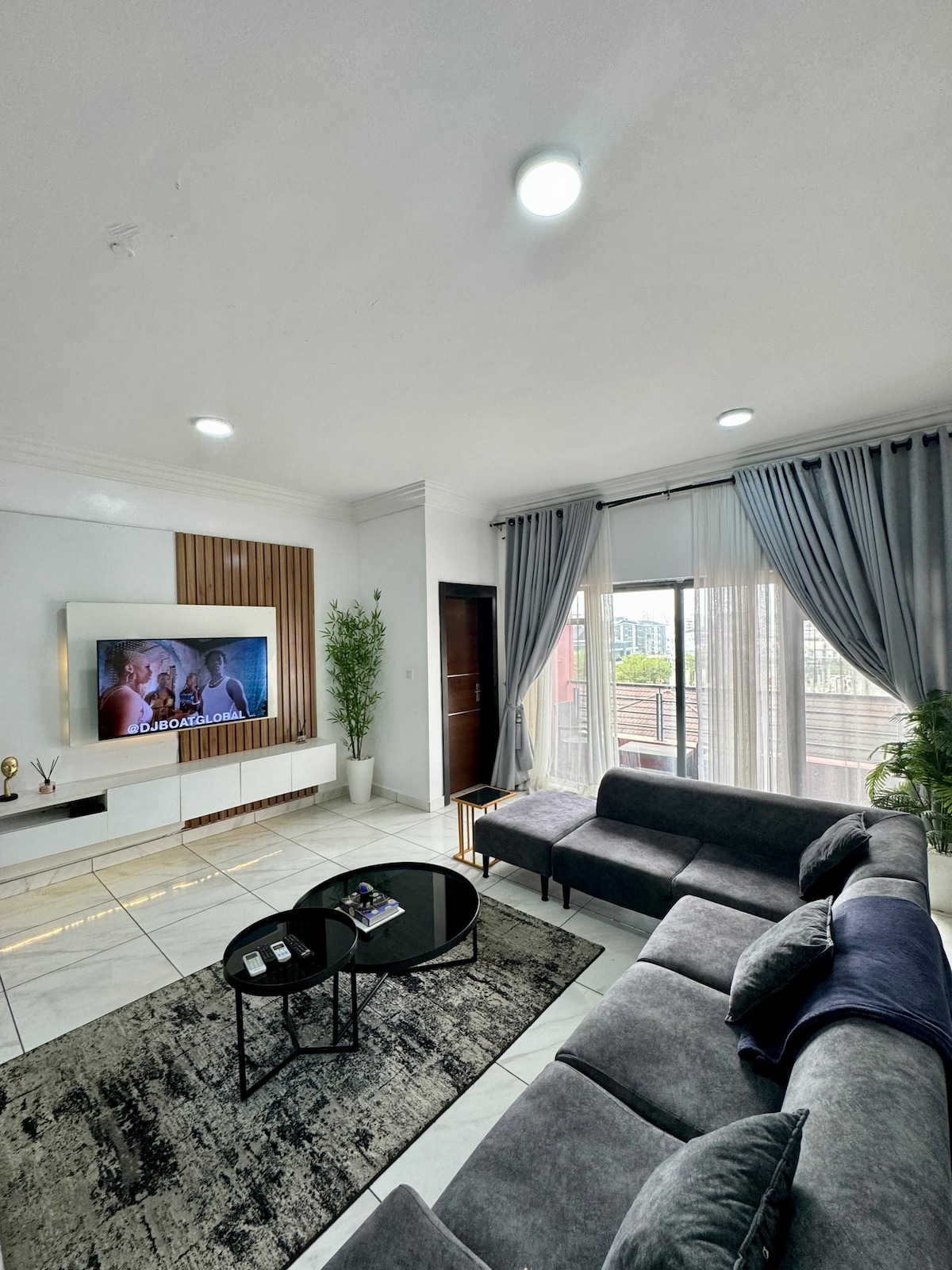 The living room area features a dark grey sectional sofa positioned for comfort. A sleek TV is mounted on the wall, accompanied by a light wood accent. Natural light filters through large windows, framed by elegant drapes. A round black coffee table sits atop a textured area rug.