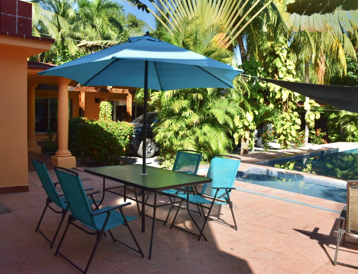 An outdoor dining area is highlighted by a large turquoise umbrella, providing shade over a table surrounded by six matching chairs. Lush green foliage frames the space, with a glimpse of a pool reflecting the sunlight in the background.