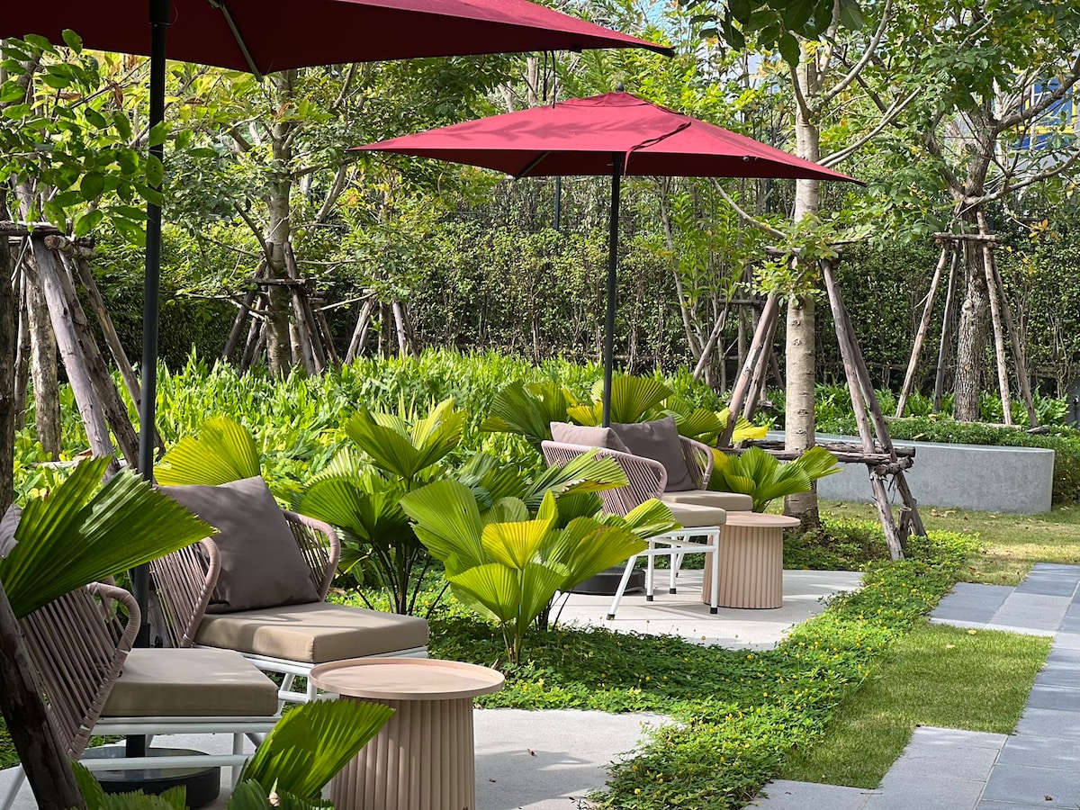 The outdoor space features a series of comfortable lounge chairs paired with small tables. Shade is provided by large maroon umbrellas, and lush greenery surrounds the area, enhancing the sense of tranquility. Various palm plants add to the serene and inviting environment.