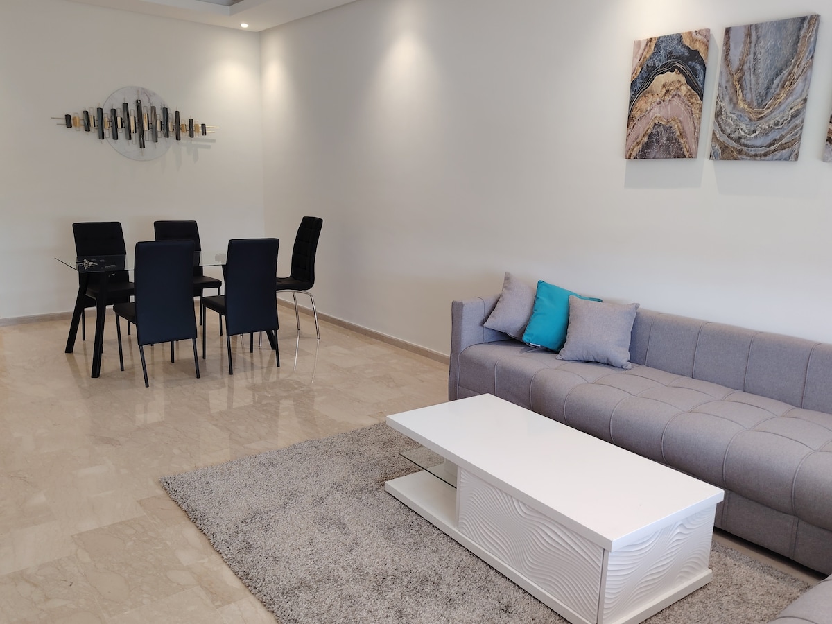 A modern living area showcases a plush gray sofa adorned with two decorative pillows. A light rug covers the tiled floor. A sleek coffee table sits in front of the sofa, while a dining table with six black chairs is positioned nearby, adding to the spacious atmosphere.