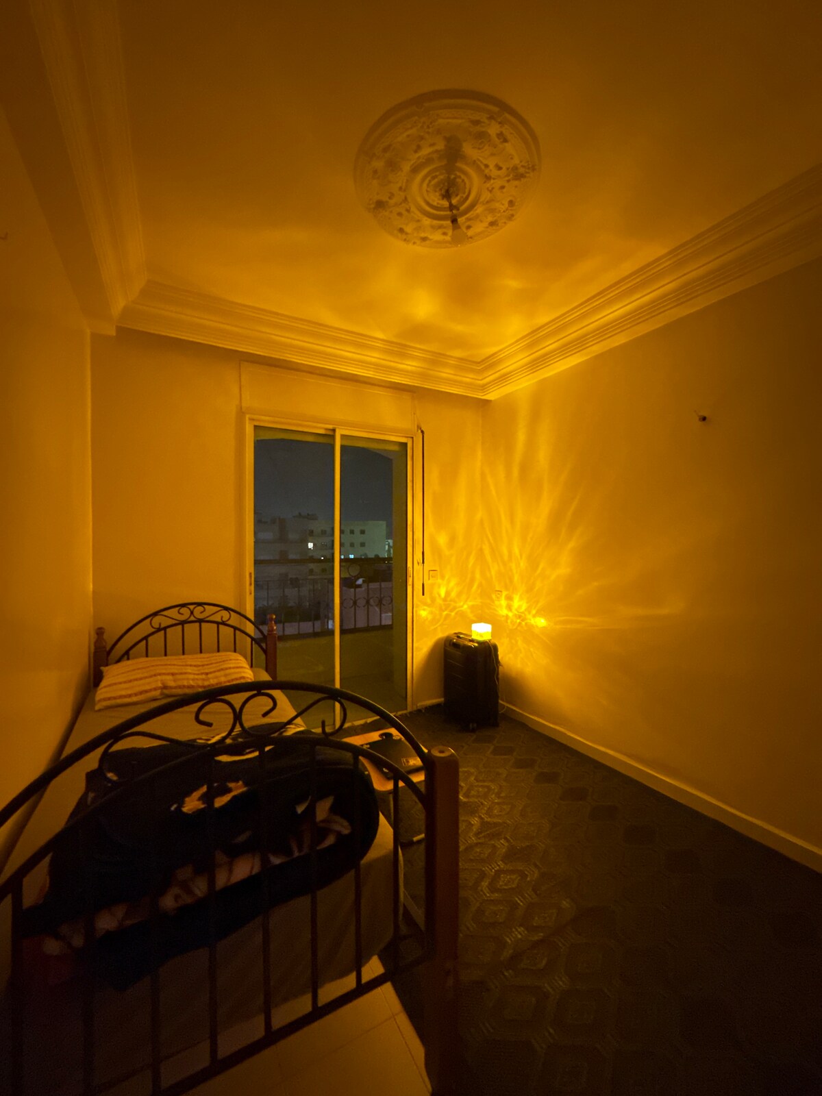 A cozy bedroom is illuminated by warm yellow tones, creating a calming atmosphere. Two twin beds are positioned near a balcony door, allowing for natural light. The room features a decorative ceiling with an ornate design, and a small lamp emits a soft glow on the floor.