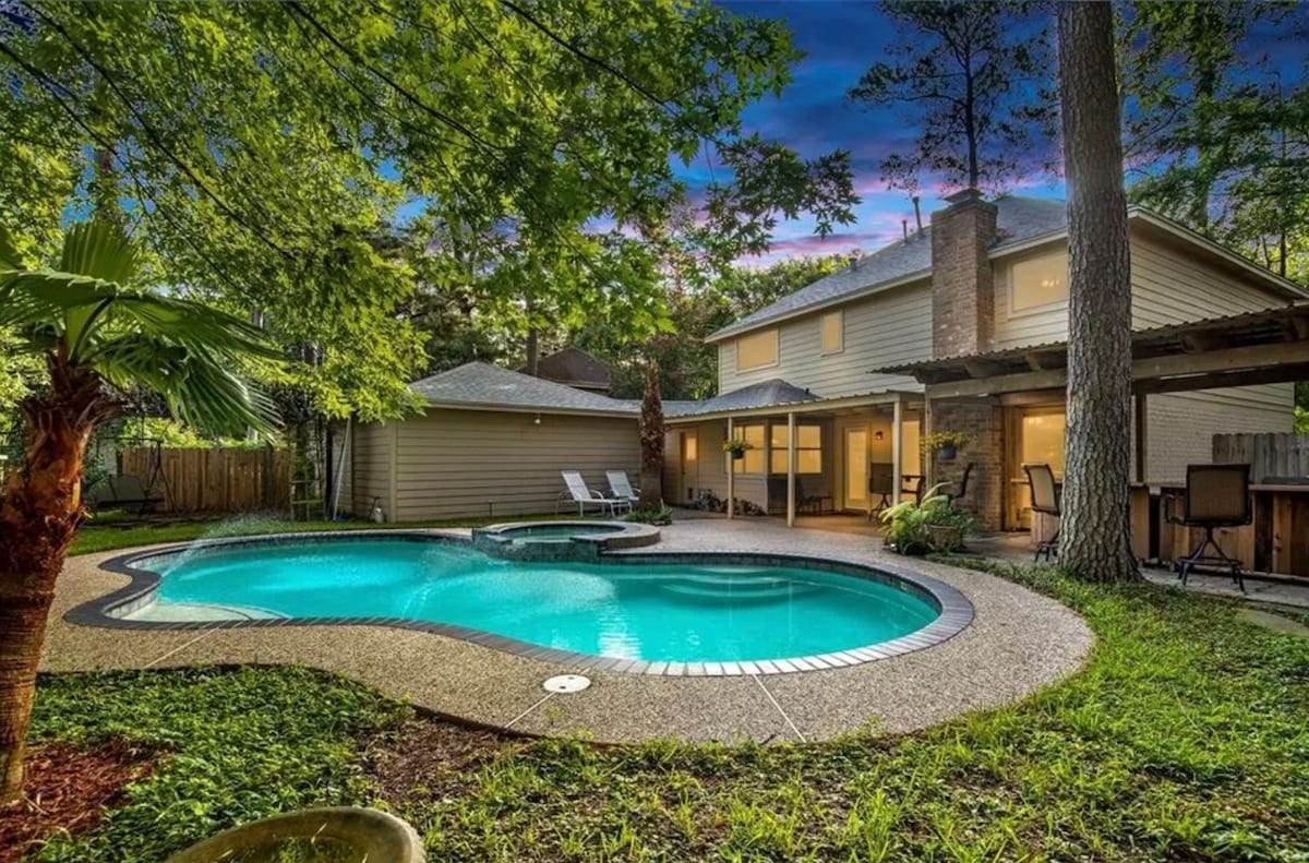 A serene backyard scene features a private pool with a built-in spa, surrounded by lush greenery and tall trees. The inviting pool area is complemented by lounge chairs, while the home stands in the background, showcasing a covered patio that provides additional outdoor space.