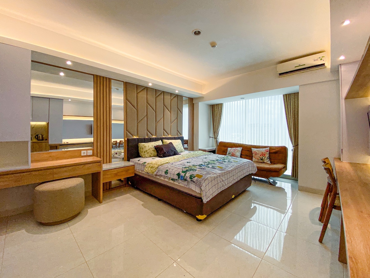 A spacious studio room is furnished with a queen bed and a sofa. The light-colored walls are complemented by large windows dressed with sheer curtains, allowing natural light to fill the space. A wooden work table and seating area are visible, enhancing the room's functionality.