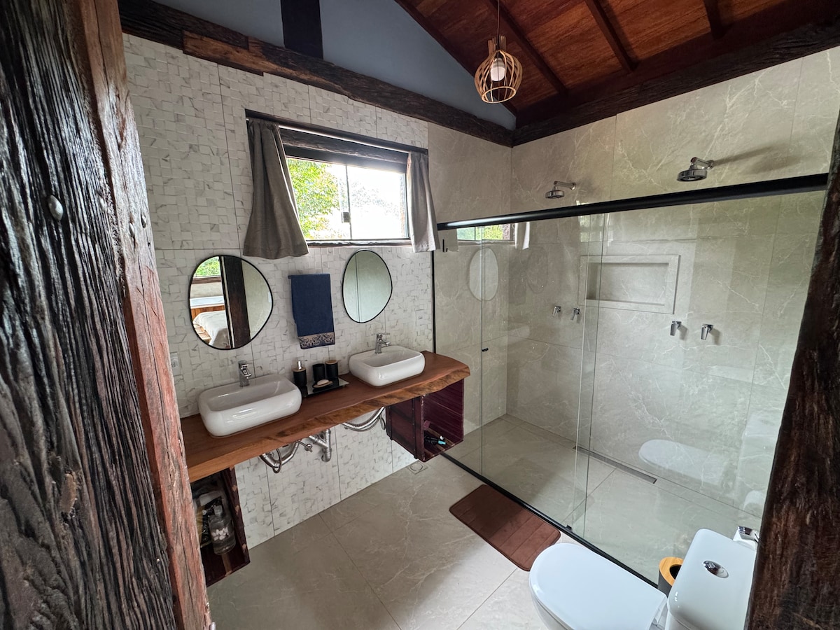 The bathroom features a double vanity with two sinks, accompanied by round mirrors adding elegance. A glass-enclosed shower area sits to the side, while light-colored tiles give a spacious feel. Natural light filters through a window, enhancing the serene atmosphere.