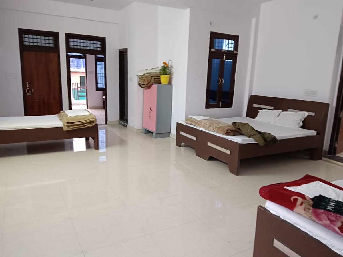A spacious room is presented with two separate beds and a neutral-toned floor. Large windows provide natural light, with two doors visible at the far end. An additional seating area includes a small cupboard, and blankets are casually placed on the beds.