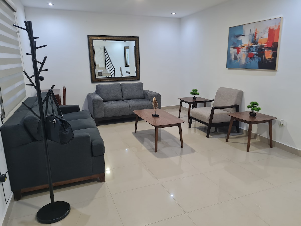 A modern living area is presented with two gray sofas and a single armchair, complemented by a wooden coffee table. Decorative elements include small plants and a wall art piece that adds a touch of color, all on a glossy tiled floor under soft lighting.