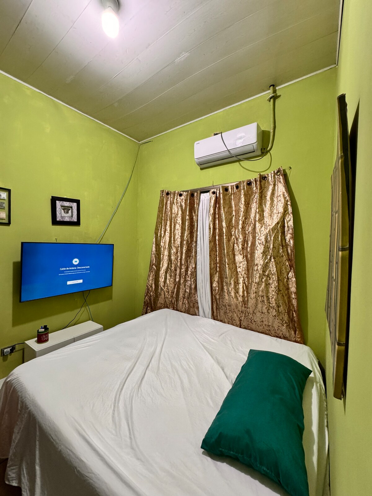 A cozy room features green walls and a spacious bed covered with a white blanket. A green accent pillow adds a touch of color. The area includes a mounted television and golden curtains framing a window, providing a sense of comfort and privacy.