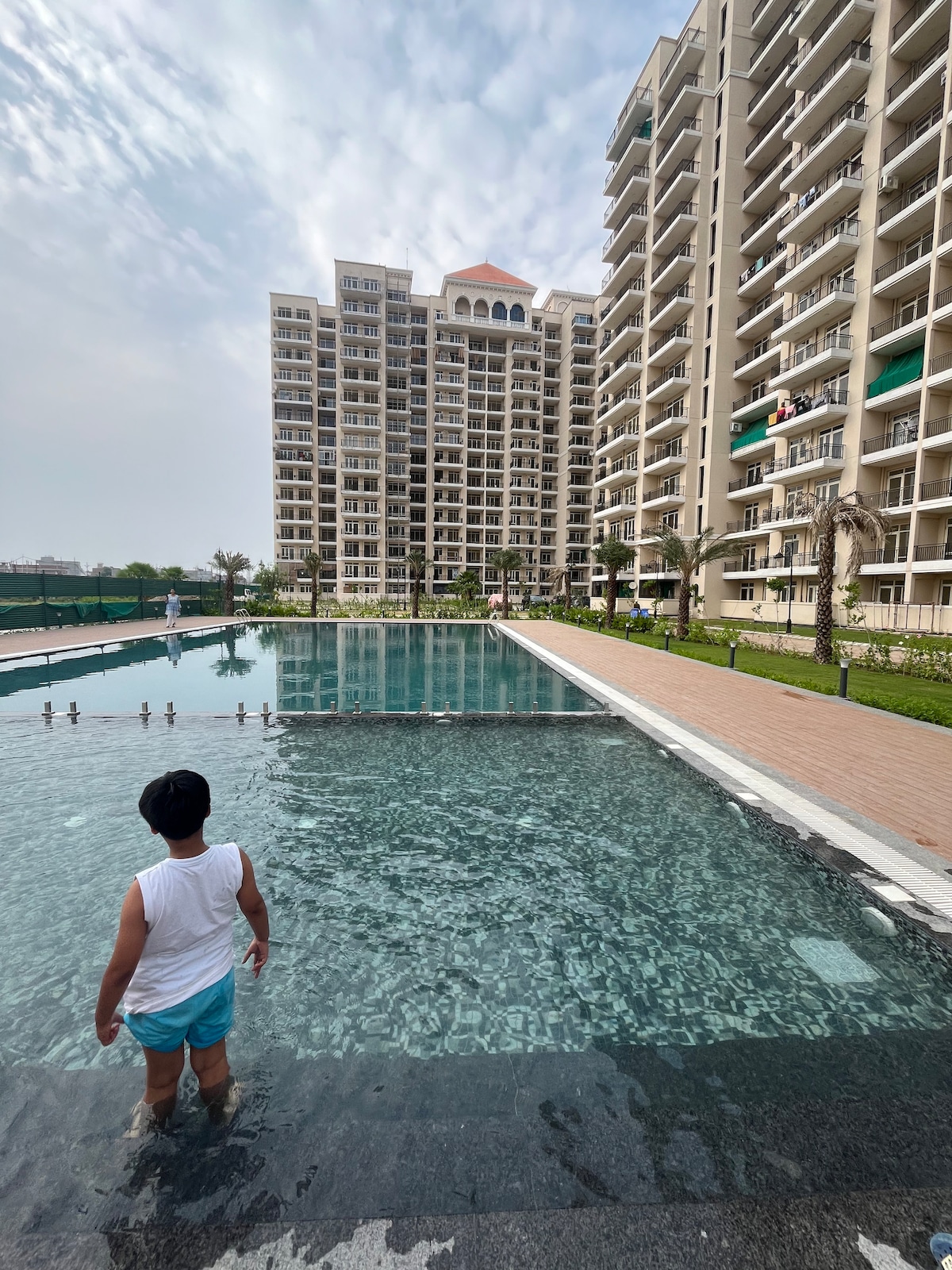 A large swimming pool is visible, reflecting the sky and the surrounding buildings. A child is standing at the pool’s edge, with water gently lapping against the sides. Modern apartment buildings surround the area, showcasing a blend of architectural styles.