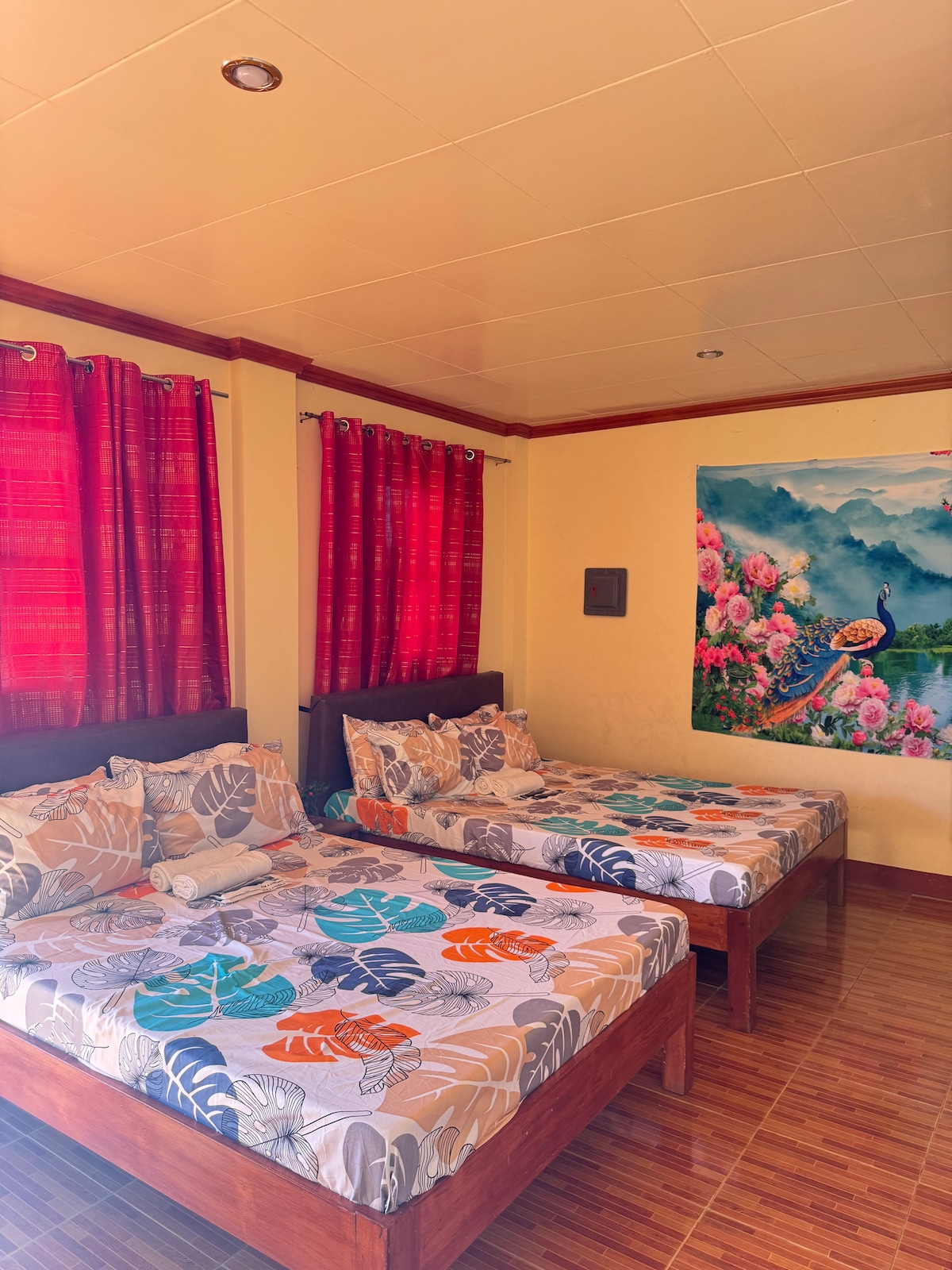 The cozy sleeping area features two spacious beds adorned with patterned bed covers reflecting vibrant floral and marine themes. Rich red curtains frame the windows, allowing natural light to enhance the room's warmth. A large mural depicting mountains and flowers adds a calming touch to the space.