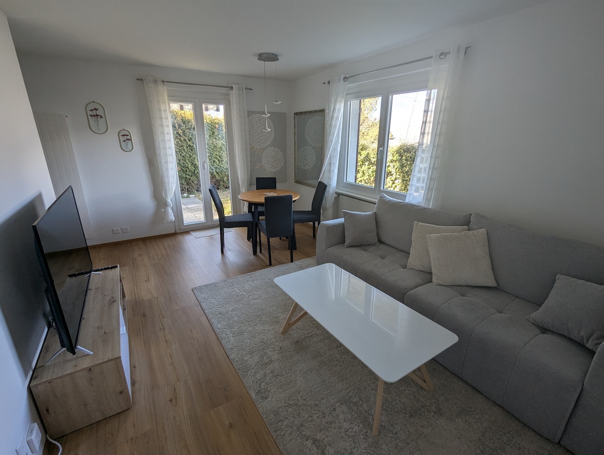 A bright living space is presented, featuring a light-colored sofa with plush cushions and a sleek coffee table. Natural light filters through the windows adorned with sheer curtains, illuminating a dining area with a round table and four chairs, set against a backdrop of greenery.