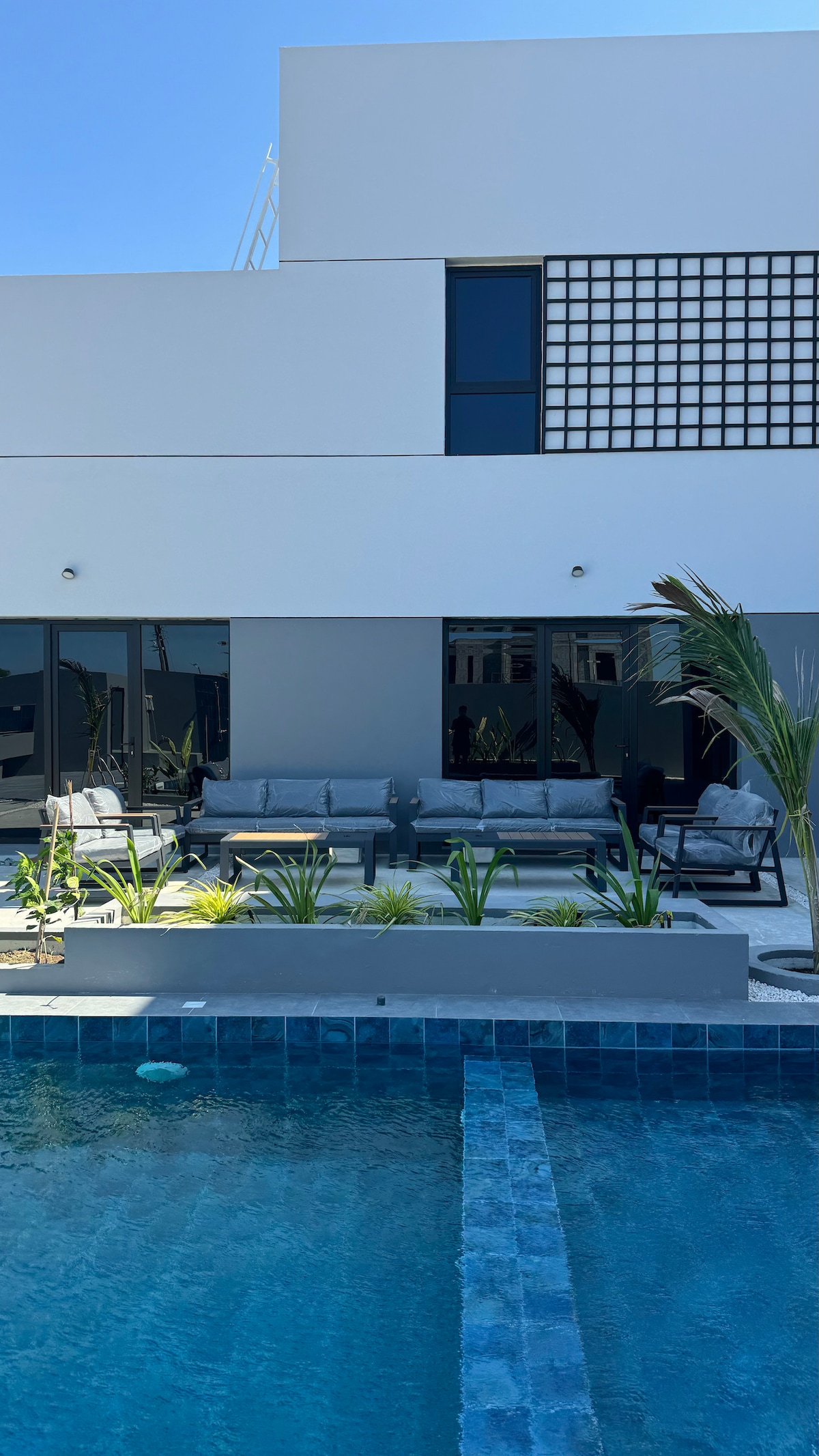 An inviting outdoor area features a swimming pool alongside a spacious lounge space. Comfortable seating arrangements accommodate multiple guests, with greenery and decorative elements enhancing the setting. Large glass doors provide views into the interior, allowing for a seamless connection between indoor and outdoor areas.