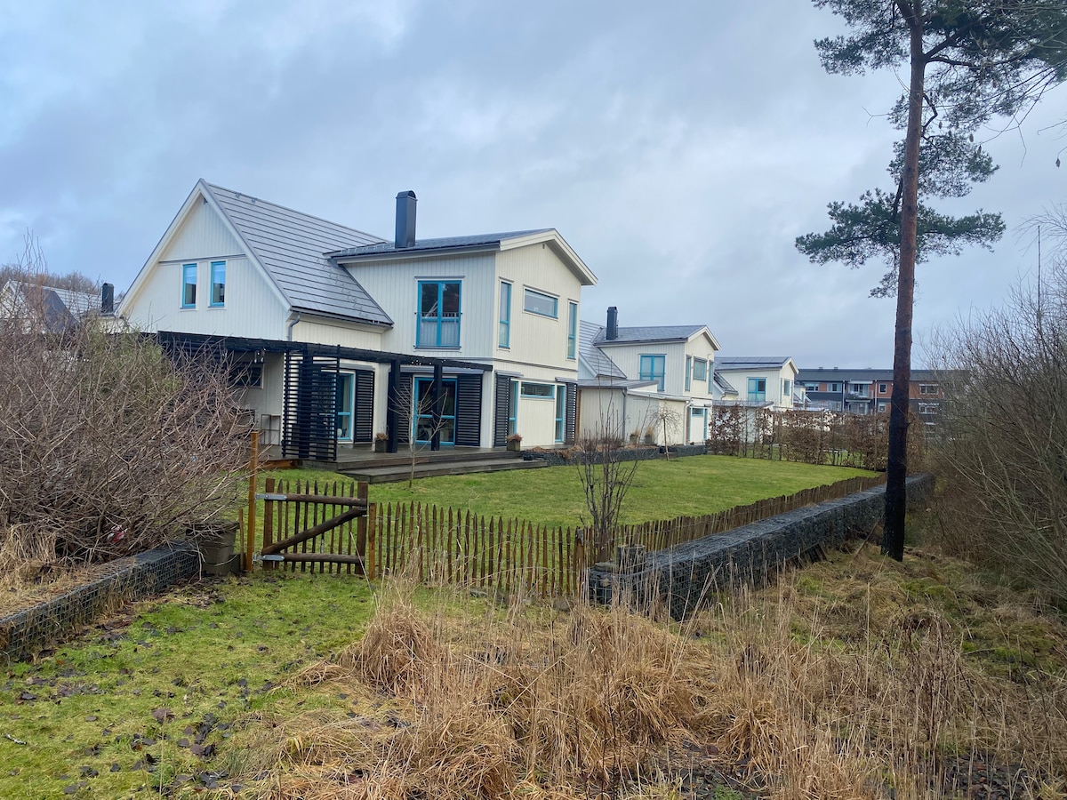 The exterior of a spacious villa is displayed, featuring a white façade with large windows. A grassy area is visible in the foreground, bordered by a wooden fence. Surrounding vegetation includes small bushes and trees, creating a natural ambiance.