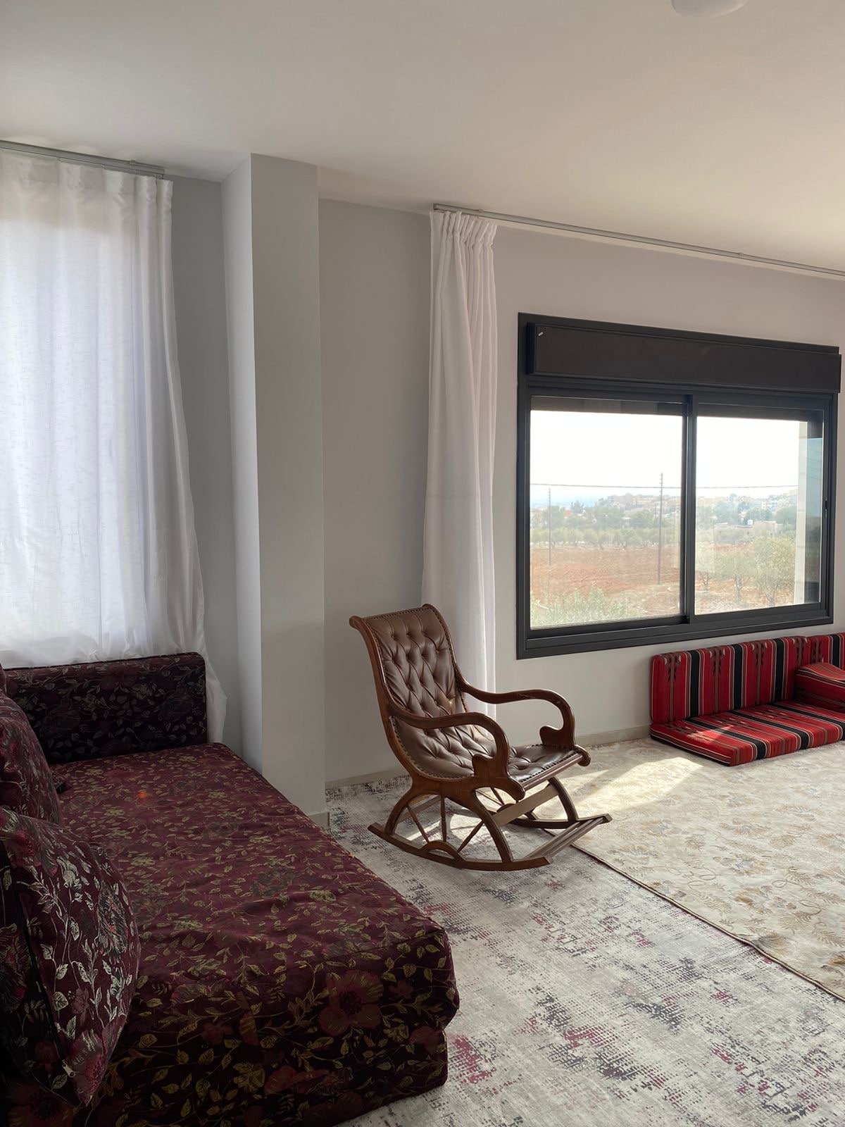 A sunlit corner features a floral-patterned sofa and a wooden rocking chair, positioned near a large window with light curtains. A soft area rug covers the floor, while additional seating is arranged on colorful cushions, creating a comfortable gathering space.