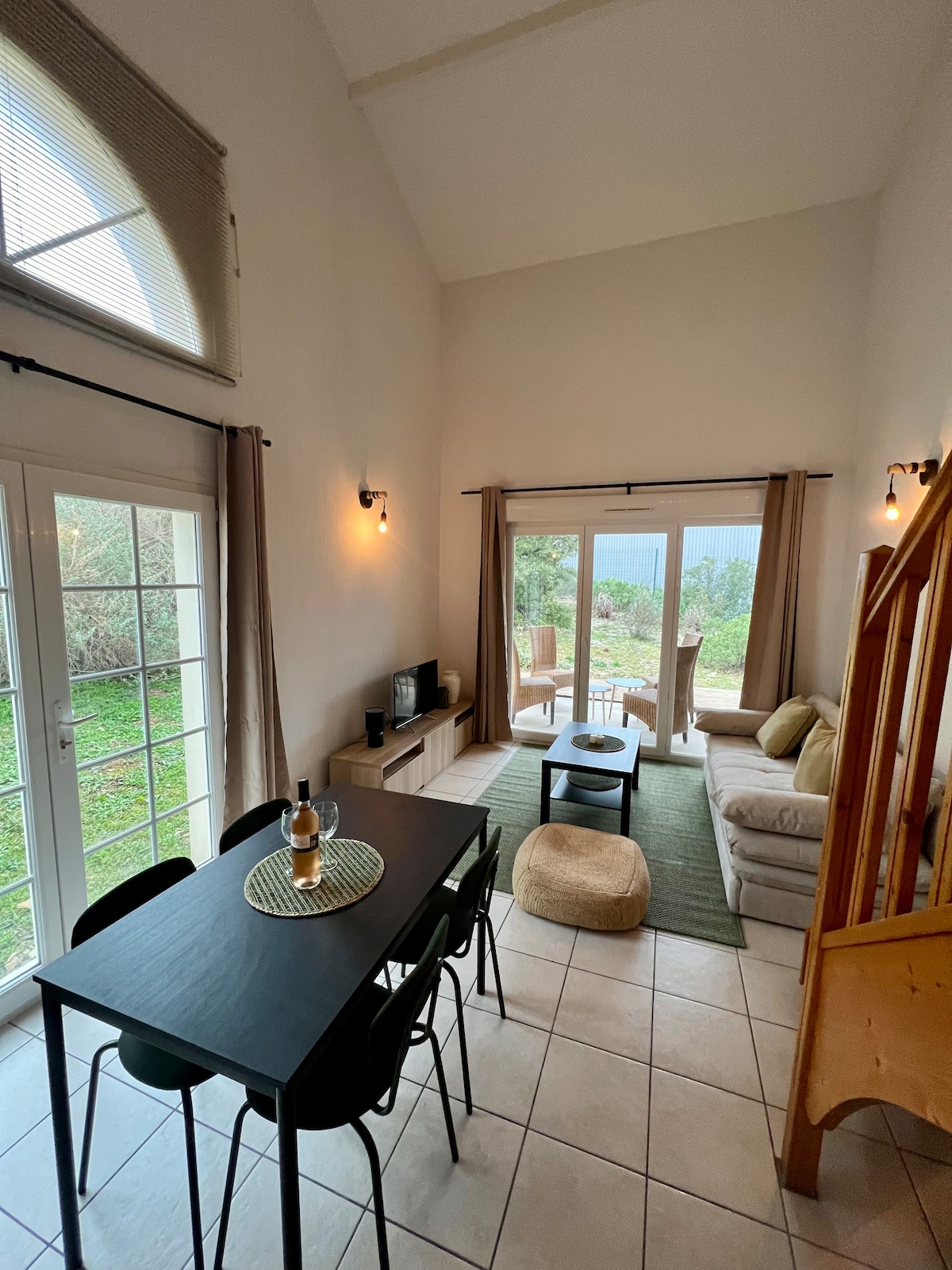 A bright living area features large glass doors leading to the outdoor space. A cozy seating arrangement includes a sofa and coffee table, alongside a dining table set for four. Natural light highlights the neutral tones and open layout, creating a welcoming environment.