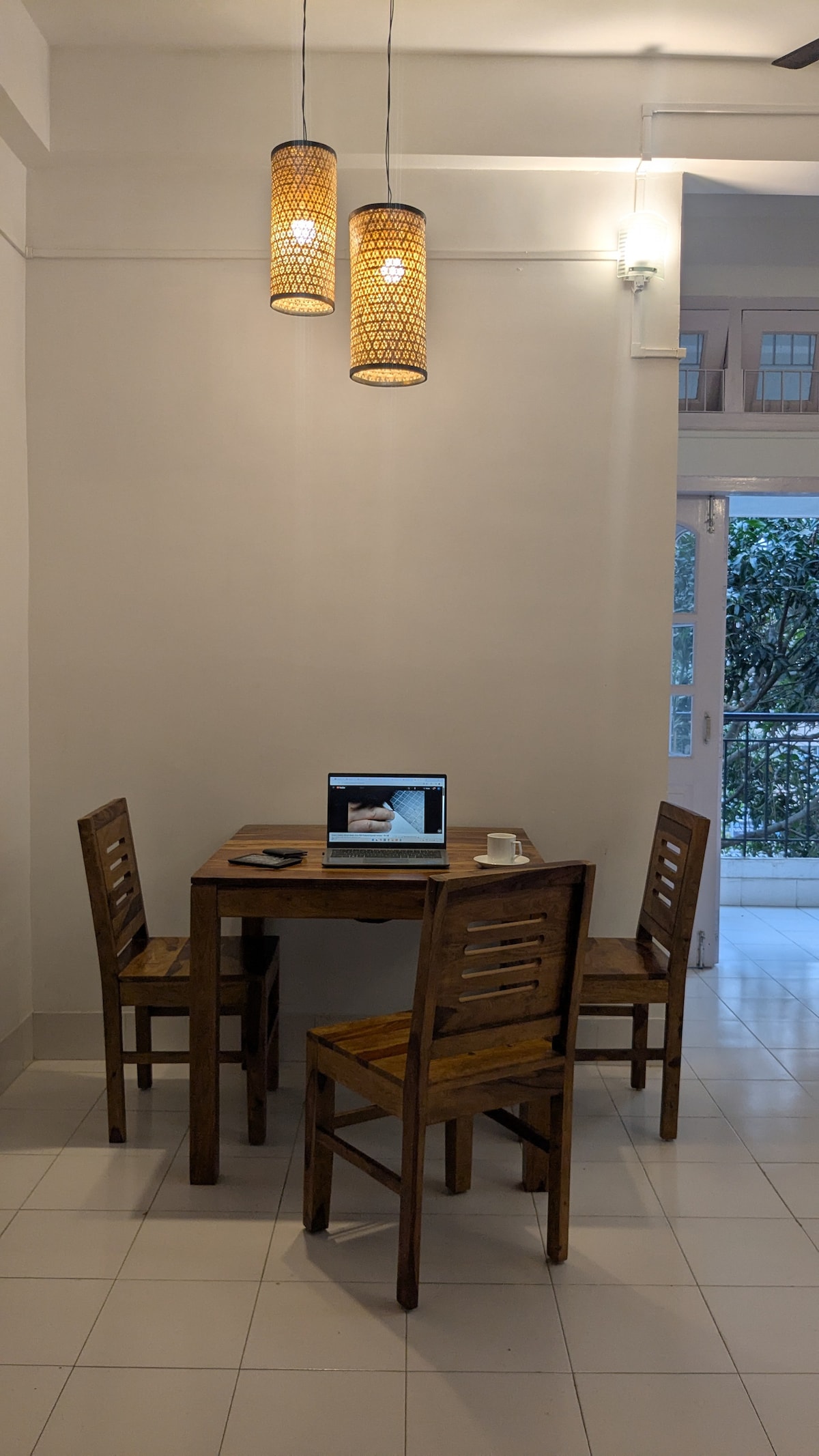 A simple wooden dining table with three matching chairs is positioned in a well-lit corner. Two pendant lights hang above the table, providing warm illumination. A laptop and a mug are placed on the table, and large windows offer a view of the outdoors.