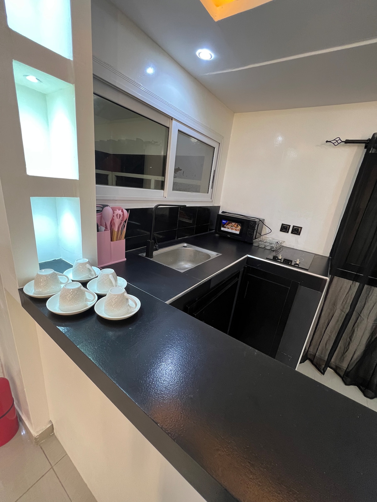 A compact kitchen area is designed with sleek black countertops and cabinetry. A stainless-steel sink is positioned under a window, allowing natural light to enter. Several white cups are neatly arranged on the countertop, alongside a microwave and utensils stored in a pink container.