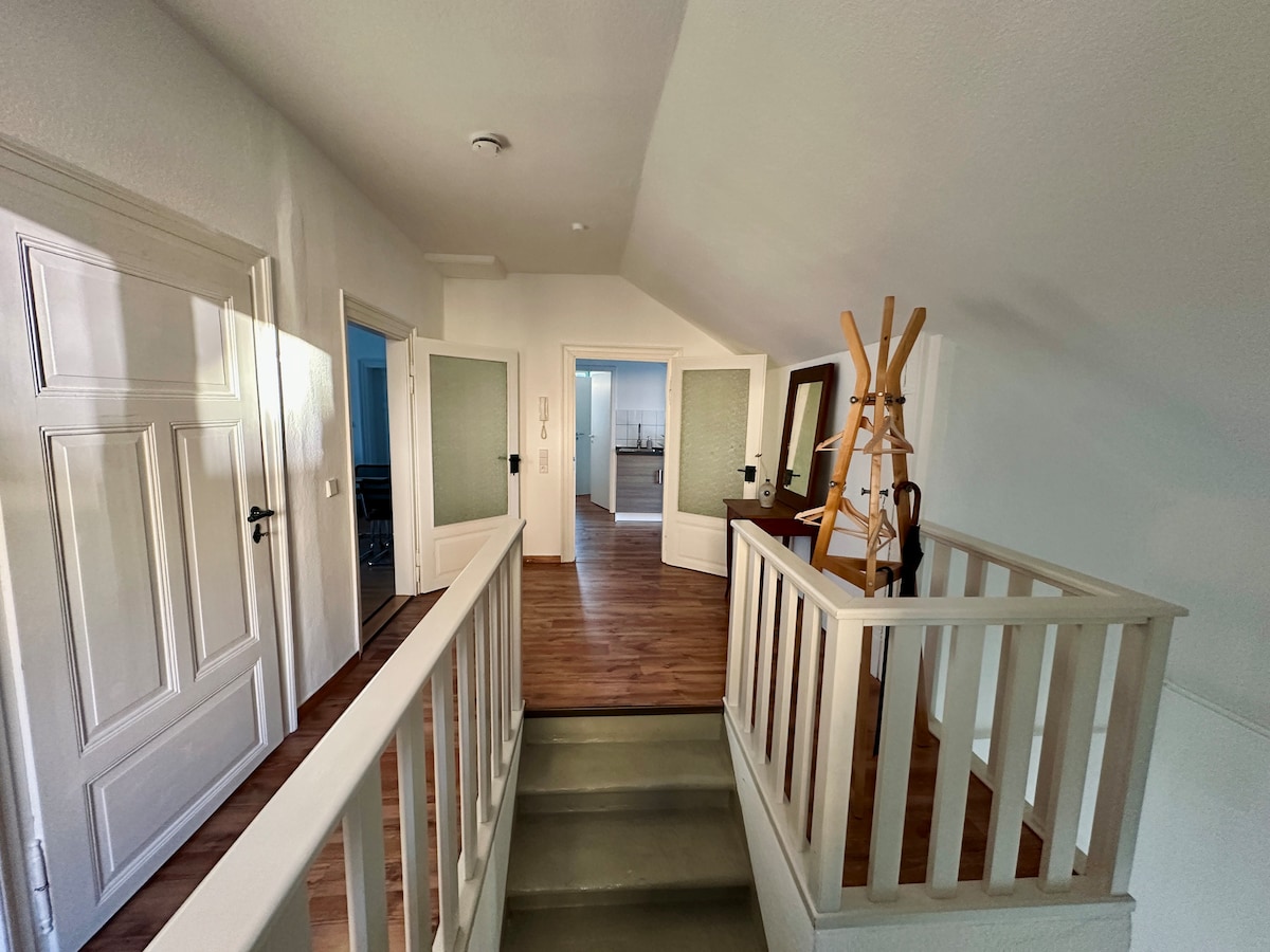 A bright hallway is visible, featuring a wooden staircase leading to a landing area. White walls create a spacious feel, while doors to the left and right offer access to other rooms. A wooden coat rack stands against the wall, complementing the hardwood floor.