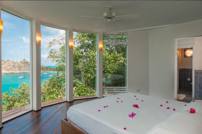 A spacious bedroom is showcased, featuring a large bed adorned with petals. Natural light pours in through circular windows, revealing scenic views of the bay and lush greenery. An attached bathroom is visible, contributing to the room's serene and private ambiance.