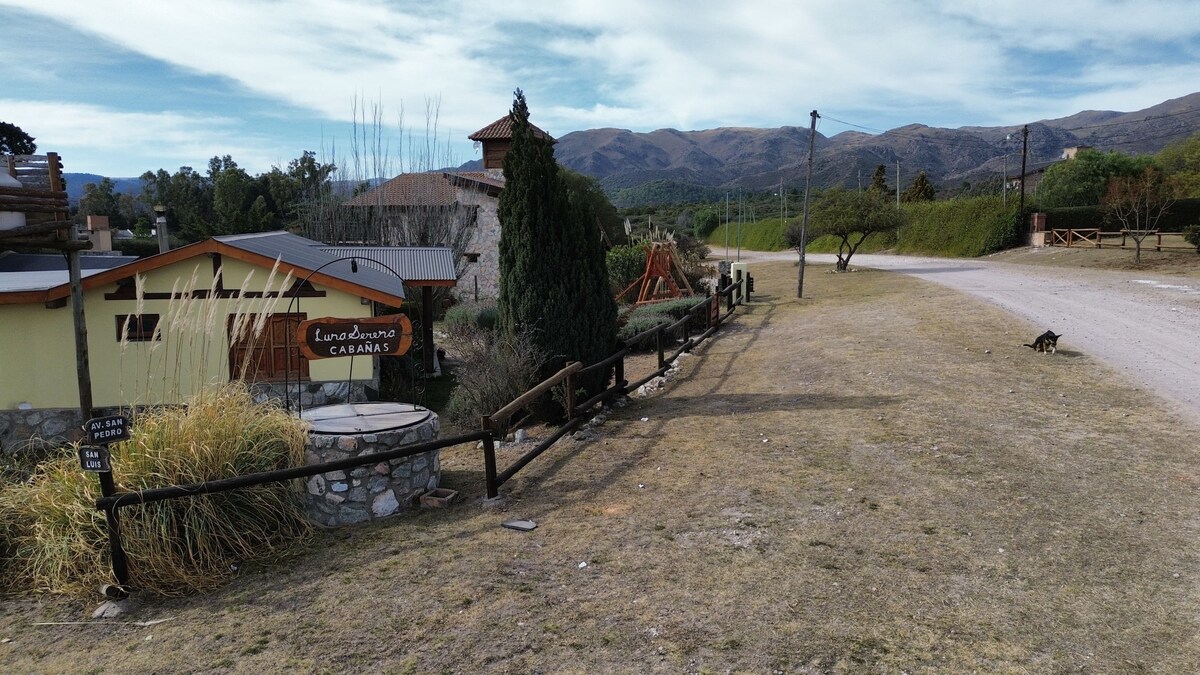 The image showcases a peaceful rural landscape with mountain views in the background. A well-maintained pathway leads past several buildings, including visible signage for the accommodation. Lush greenery and a small cat can be seen along the dirt road, creating a tranquil environment.
