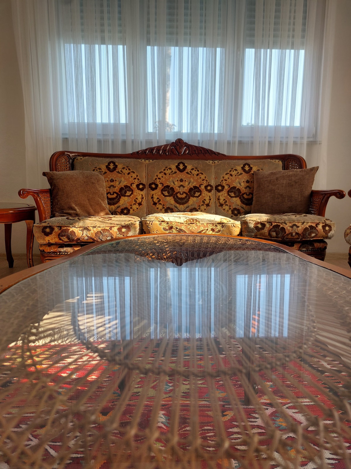 A vintage-style sofa with intricate floral patterns and plush cushions faces a glass-topped coffee table. Natural light filters through sheer curtains, casting soft reflections on the table's surface, while a warm wood accent table is positioned nearby.