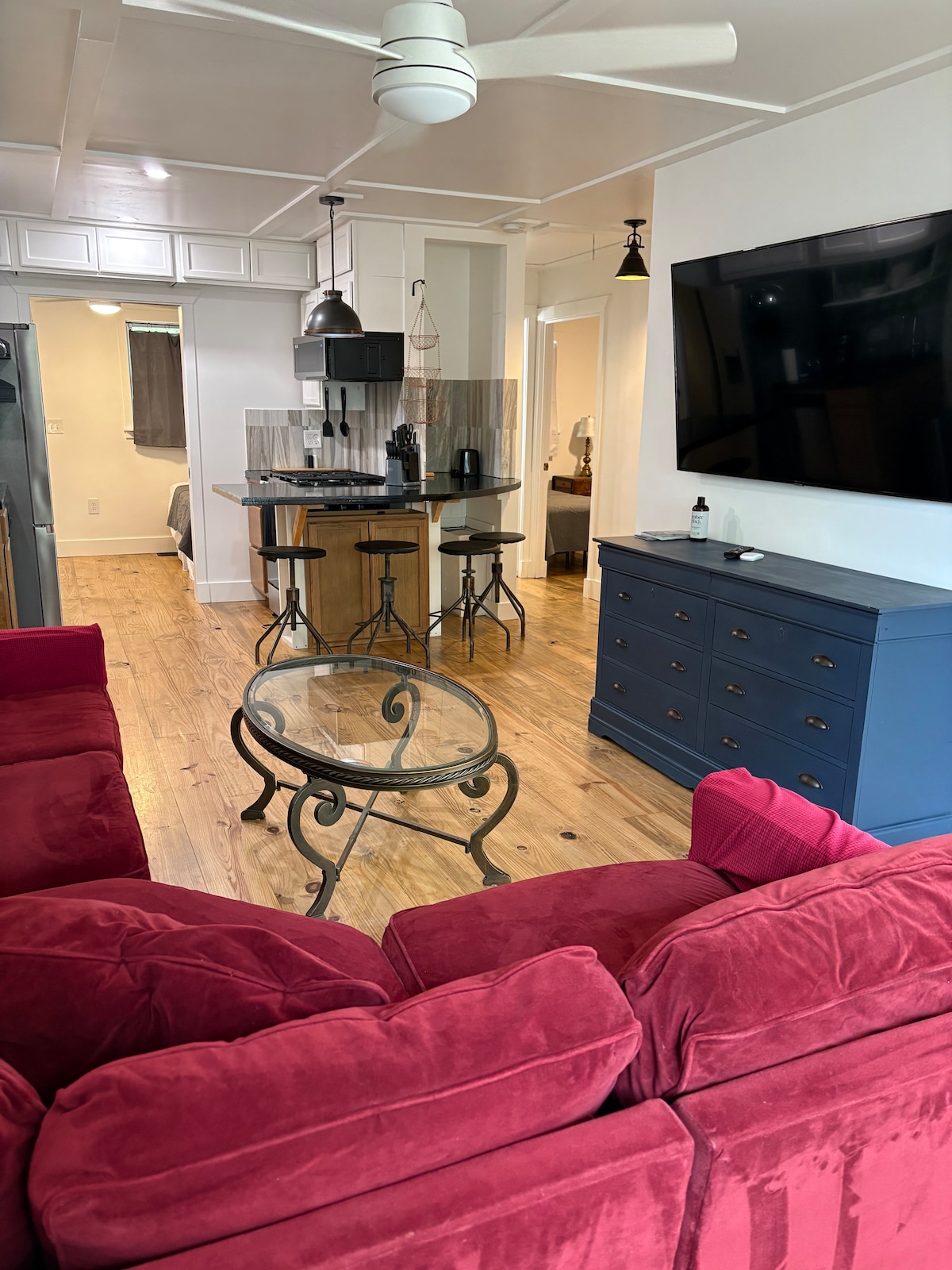 A cozy living area features a plush red sofa facing a glass coffee table with an intricate metal base. In the background, a kitchen island with seating is visible, along with a flat-screen TV mounted on the wall and warm wood flooring throughout.
