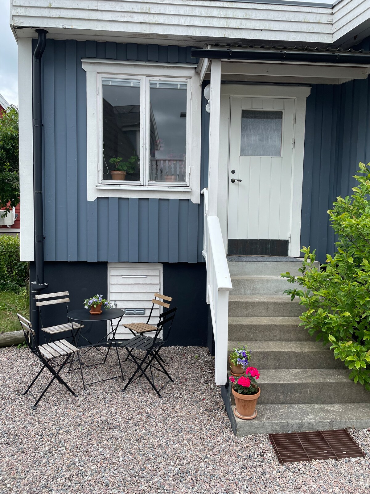 An entrance to the guest house is shown, featuring a set of two black metal chairs and a small round table placed on gravel. Potted flowers add a touch of color, and a staircase leads to the front door, which is framed by a blue-colored wooden facade.