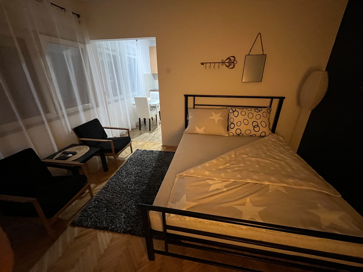 The sleep area features a bed dressed in star-patterned bedding, accompanied by a decorative pillow. A small rug lies underfoot, while two black chairs are positioned nearby. Soft light filters through sheer curtains, illuminating the inviting space.