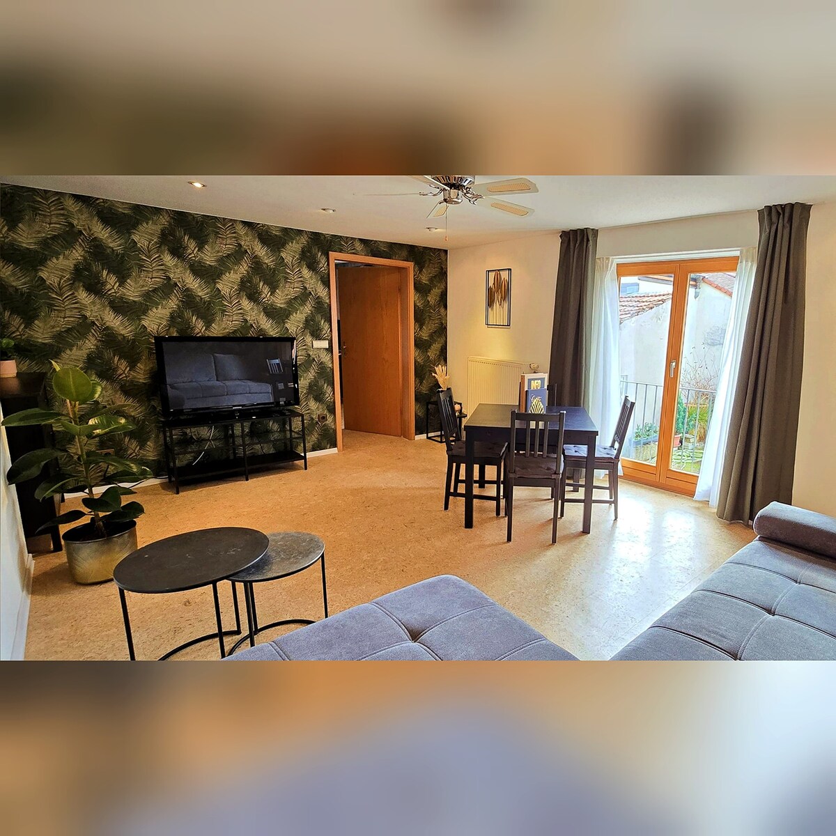 A stylish living area is presented, featuring a large television mounted on a stand. A dark table is surrounded by four chairs, and a cozy couch is visible in the foreground. Natural light enters through double doors that lead to an outdoor space, complemented by a plant in the corner.