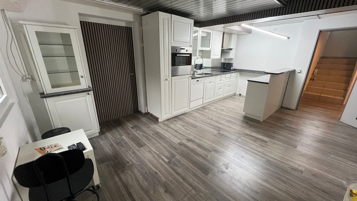 A modern kitchen space is presented with white cabinetry and sleek countertops. An oven and microwave are integrated into the cabinetry. A small dining area is shown with a dark chair at a table, while a doorway leads to a staircase visible in the background.