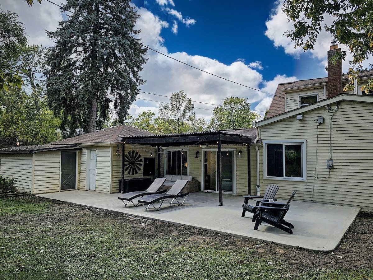 A spacious outdoor patio features a covered pergola and two lounge chairs, set against a backdrop of lush greenery. The area is designed for relaxation, with a comfortable Adirondack chair positioned nearby, perfect for enjoying the serene surroundings.