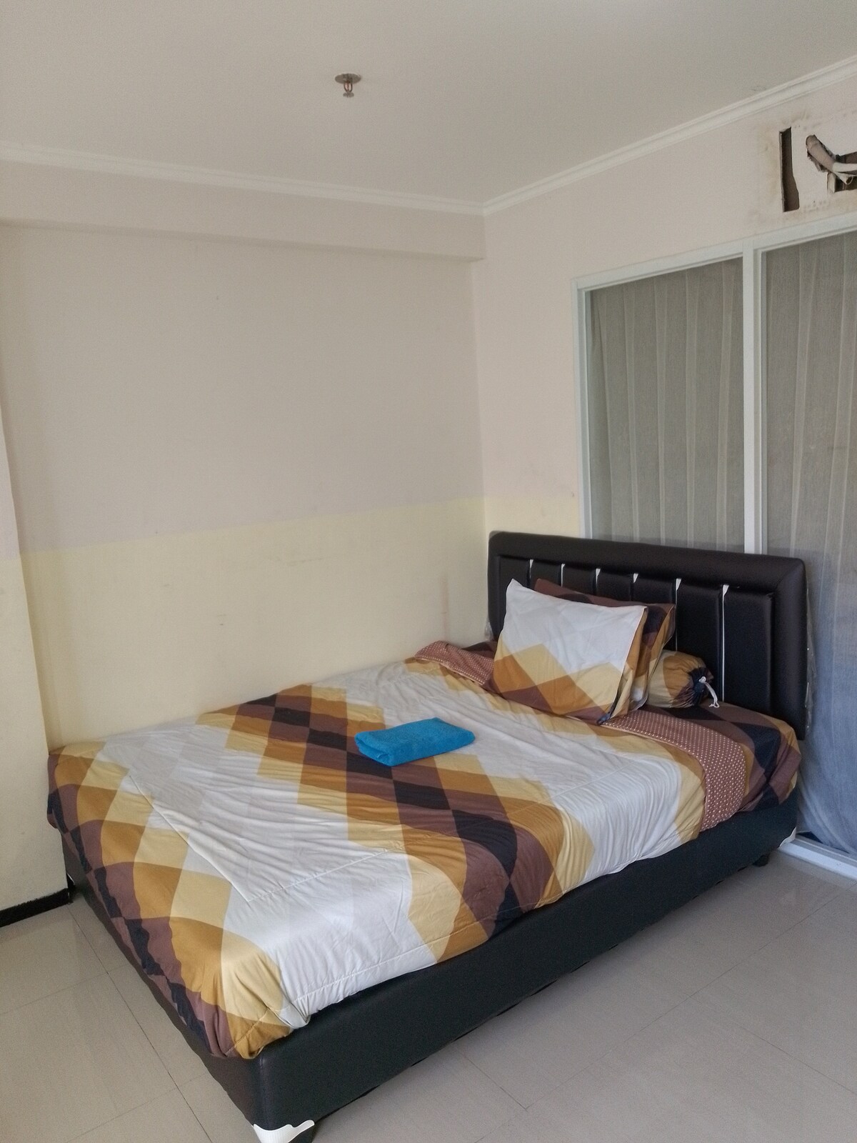 A simple sleeping area features a bed with a geometric patterned duvet and a single blue towel placed atop. Natural light is allowed in through a partially covered glass door, illuminating the neutral-toned walls and clean tile flooring.
