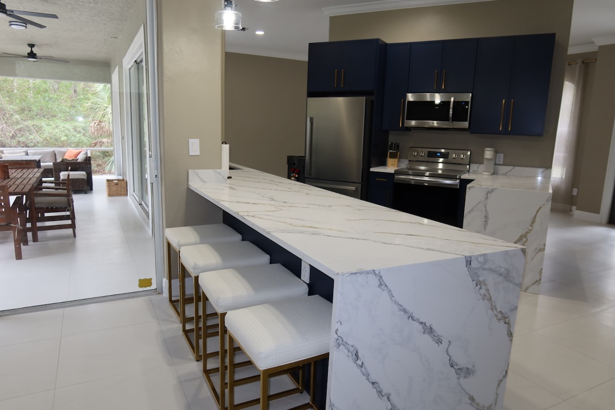 A modern kitchen space is highlighted with navy blue cabinets and stainless steel appliances. A marble island with four cushioned bar stools offers a casual dining area. Natural light streams in from large sliding glass doors leading to an outdoor seating area.
