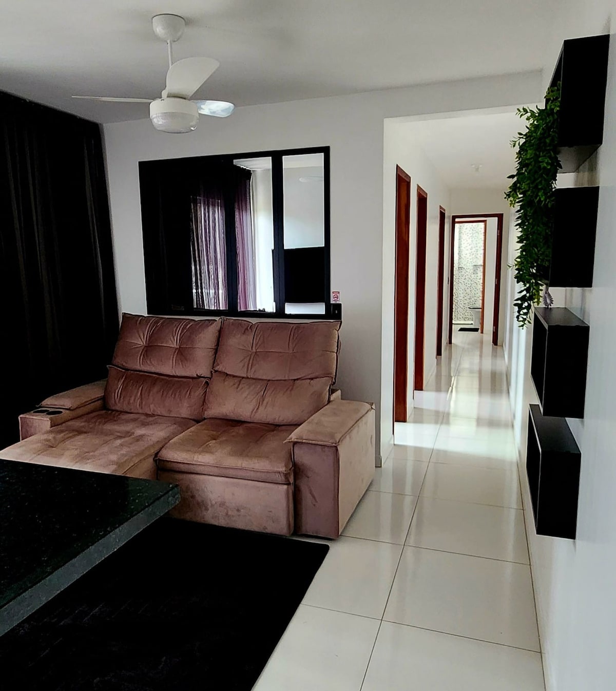 A comfortable seating area is featured, highlighted by a plush sofa facing a sleek black table. A modern ceiling fan is visible above. The hallway beyond leads to additional rooms, with contemporary decor and neutral tones evident throughout.