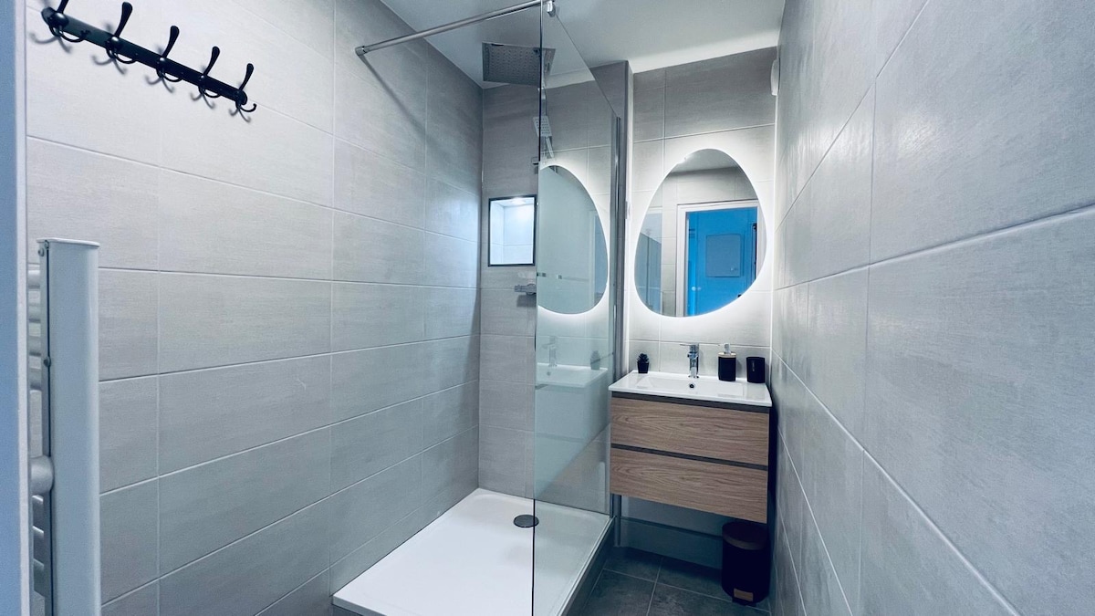A modern bathroom features a walk-in shower enclosed in glass, complemented by a wooden vanity with a round mirror above. The space is designed with neutral tiles, and there is a towel rack on the wall, creating a clean and functional environment.