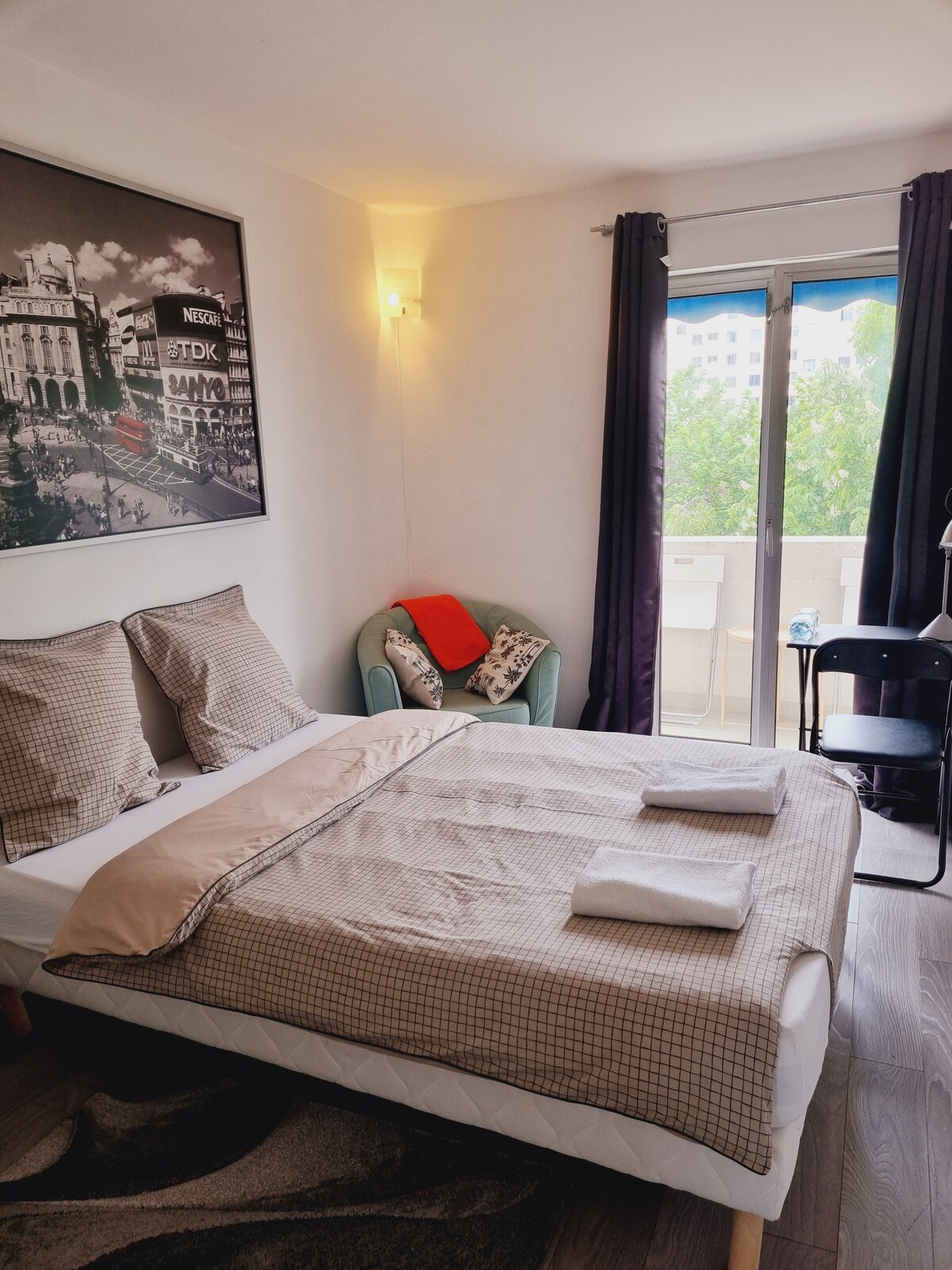 A well-lit bedroom features a spacious bed dressed in neutral-toned linens and decorative pillows. A modern armchair with patterned accents sits nearby, and a small balcony is visible through glass doors, framed by sheer curtains and dark drapes.