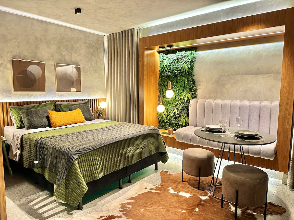 A thoughtfully arranged sleeping area features a spacious bed with a green and gray bedding set. Natural elements are incorporated, with a vertical garden beside a cozy seating nook. A modern dining table with two stools is set for two, creating an intimate dining experience.