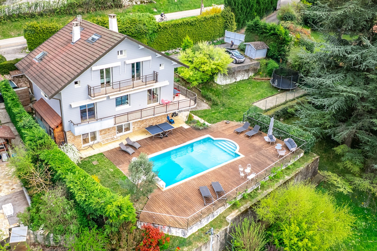 A spacious villa is surrounded by greenery, featuring a well-maintained garden and a private salt-water pool. Several sun loungers are arranged around the pool deck, inviting relaxation. The property includes a trampoline and outdoor furniture, making it suitable for family gatherings.