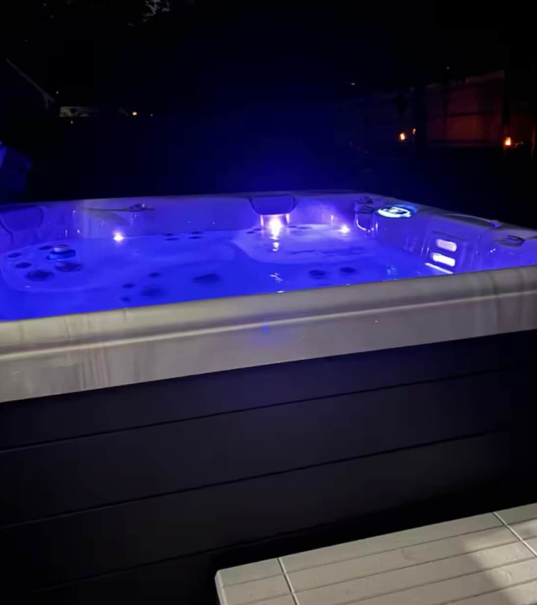 A private hot tub is illuminated by soft blue lights, creating a serene ambiance for relaxation. The sleek design features comfortable seating and various jets. A wooden platform is positioned nearby, enhancing the outdoor space.
