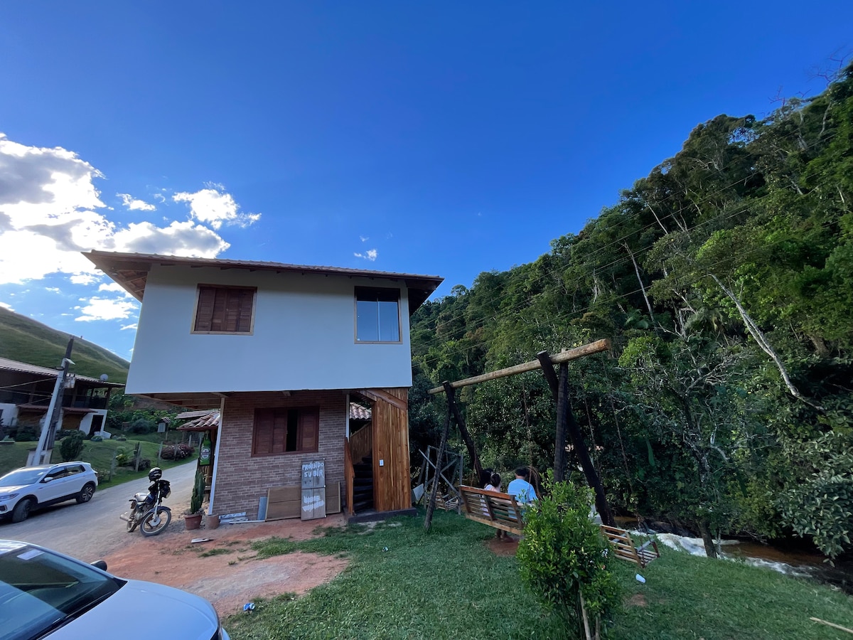 The exterior of the two-story building is visible, surrounded by lush greenery. A wooden swing is positioned nearby, offering a natural seating area. A serene river can be seen at the base of the structure, enhancing the calming nature of the surroundings.