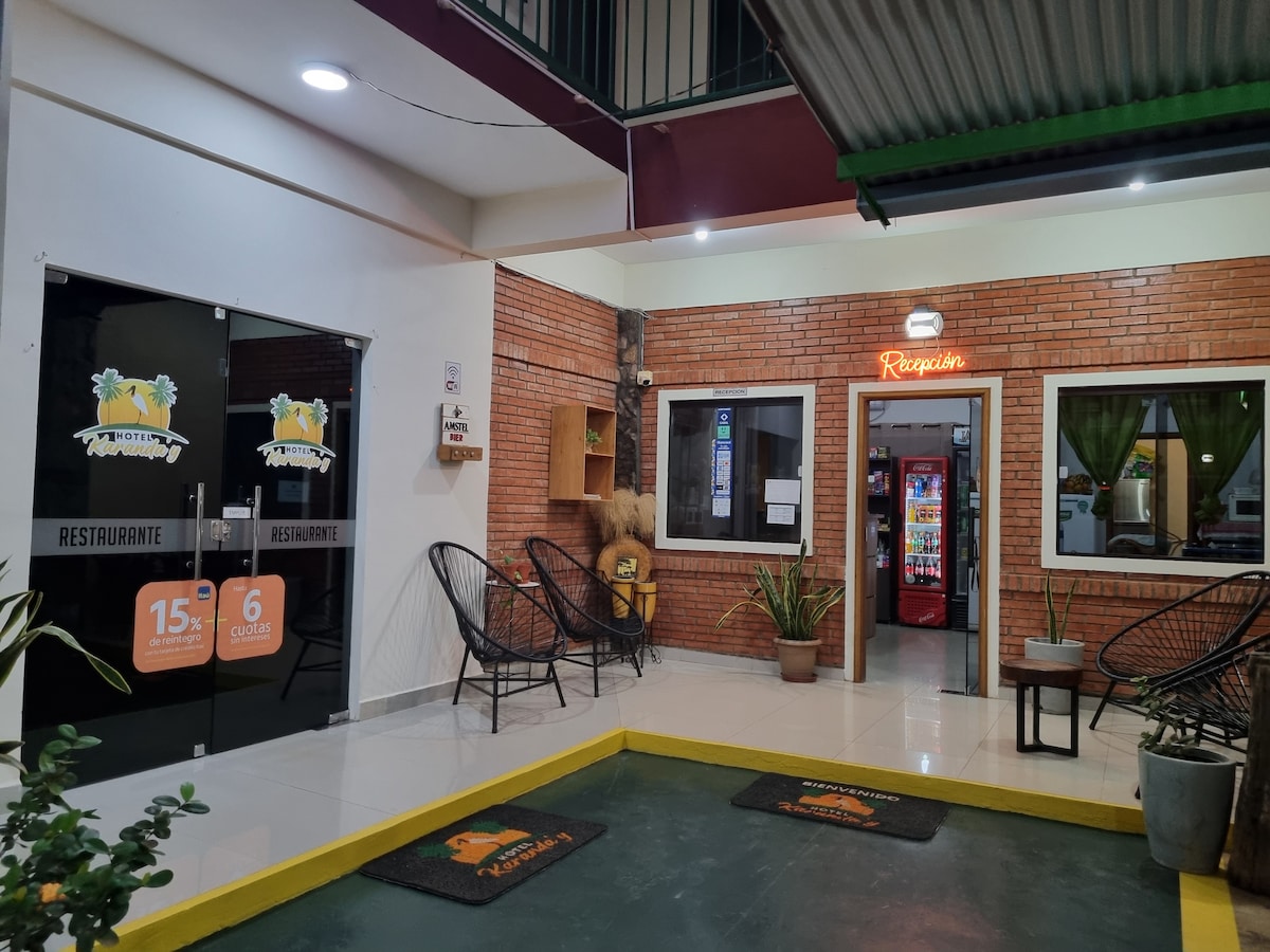 An inviting entrance showcases a modern restaurant area with brick walls and sleek glass doors. Directly visible is a seating area featuring black wire chairs. A vending machine and potted plants add to the welcoming ambiance, illuminated by soft overhead lighting.