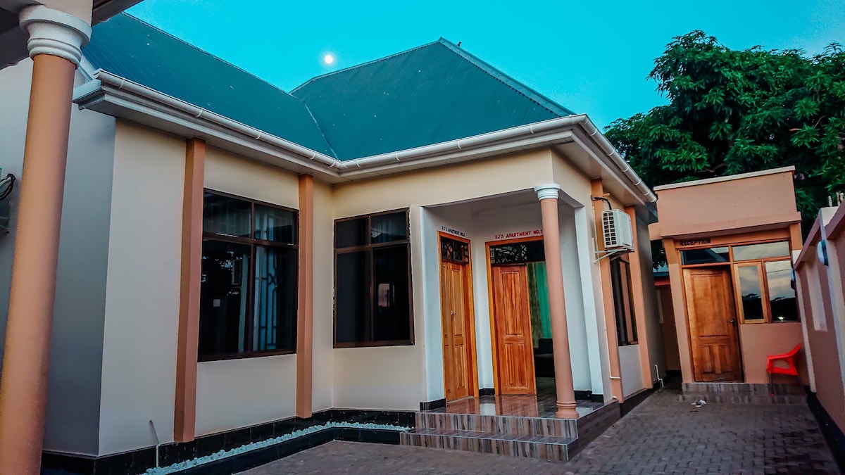 The exterior of the building features a modern architectural design with a sloped roof and multiple large windows. Wooden doors provide access to the entrance and side areas, with neatly arranged stone details along the front walkway. Lush greenery is visible in the background.