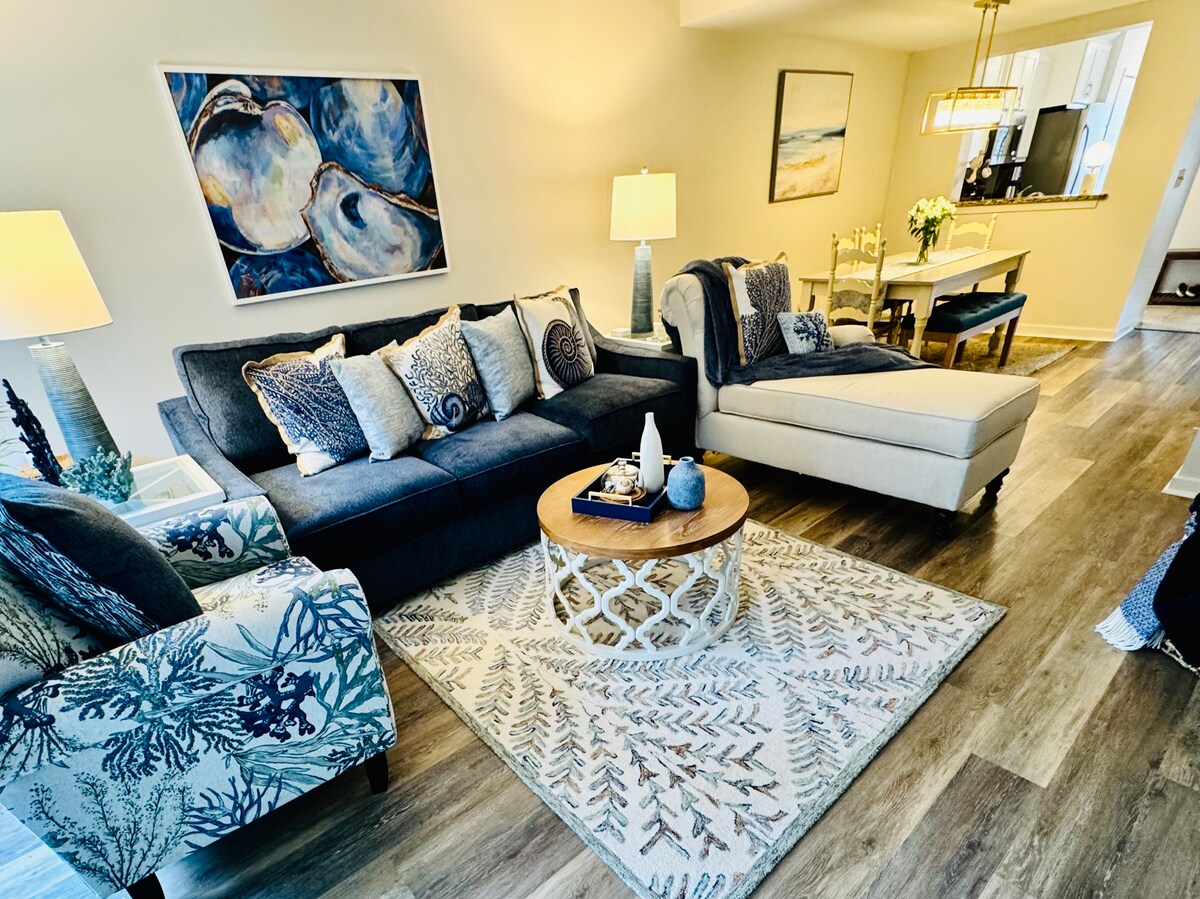 The living area features a comfortable grey sofa adorned with decorative pillows, complemented by an armchair with a blue and white marine-themed pattern. A round coffee table sits atop a patterned area rug, while a dining space is visible in the background, illuminated by natural light.