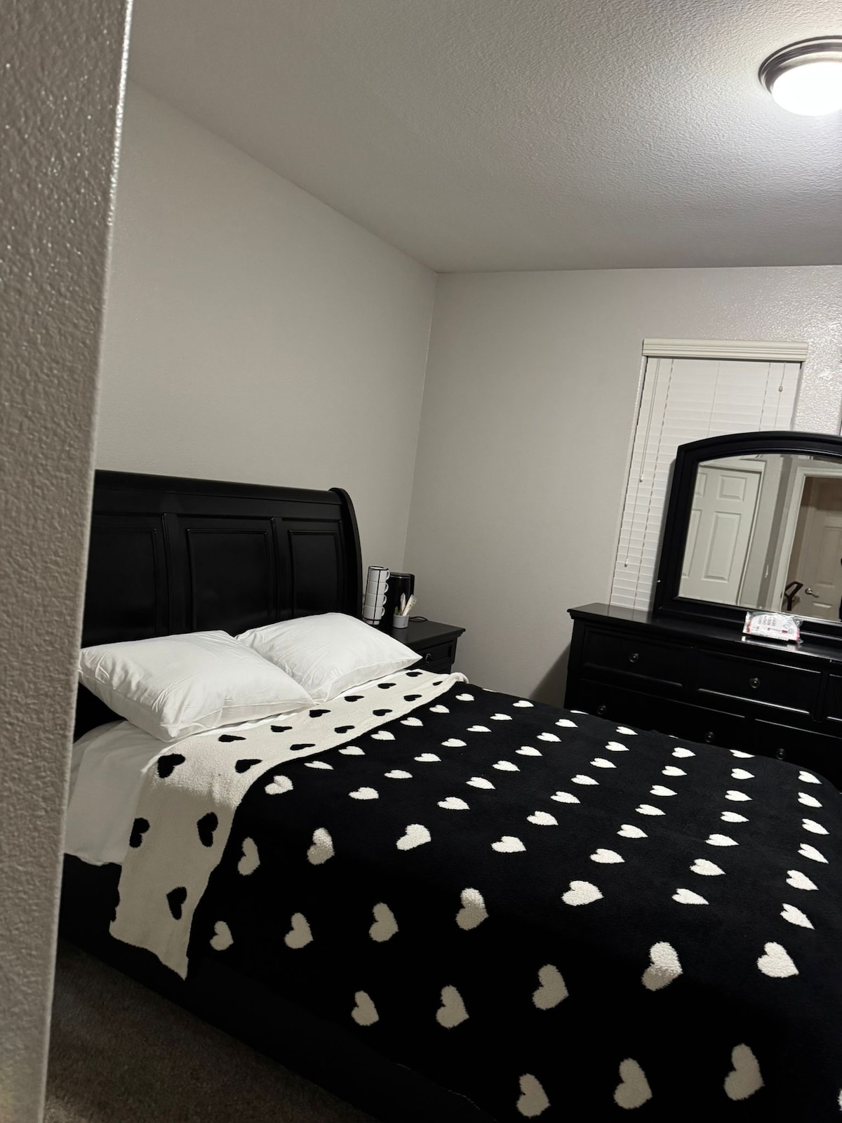 A comfortable bedroom features a dark wooden headboard and a spacious bed adorned with a black and white heart-patterned blanket. A dresser and mirror are positioned against the wall, while soft light enters through a window dressed with blinds.