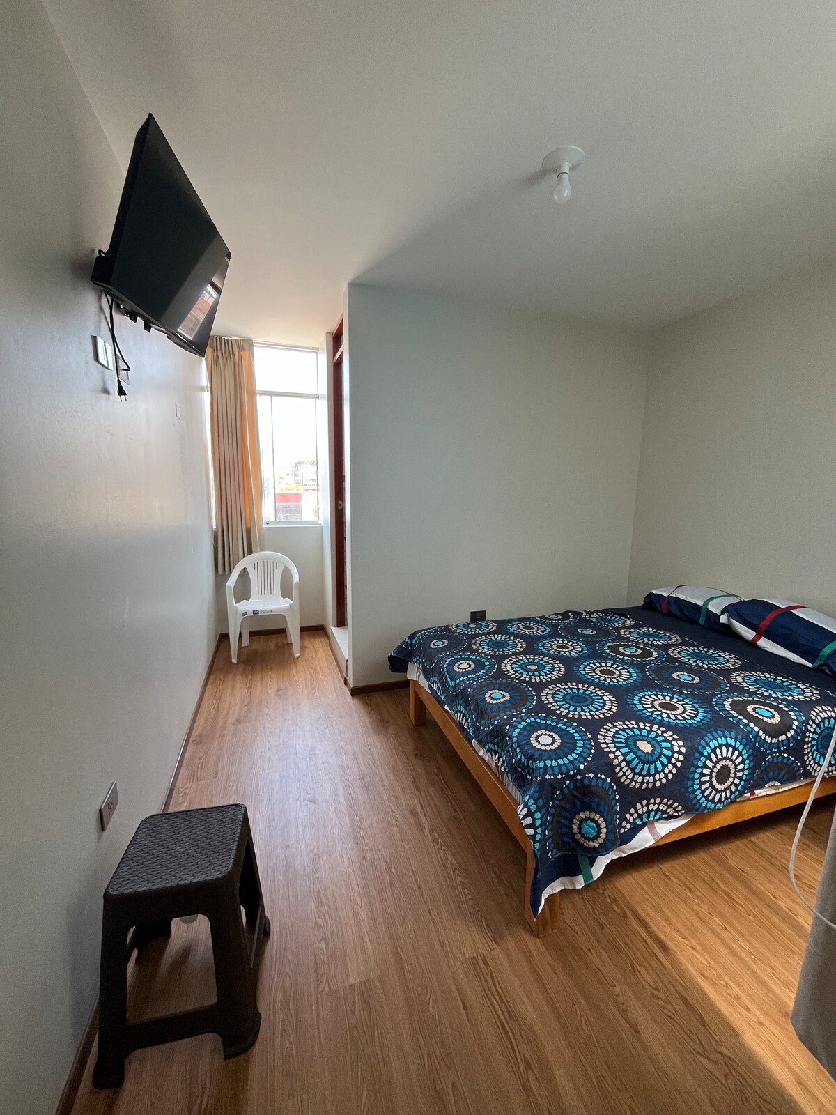 A bright bedroom features a spacious bed covered with a blue patterned blanket. A small stool is positioned beside the bed, while a white chair is located in the corner. Large windows allow natural light to illuminate the wood flooring.