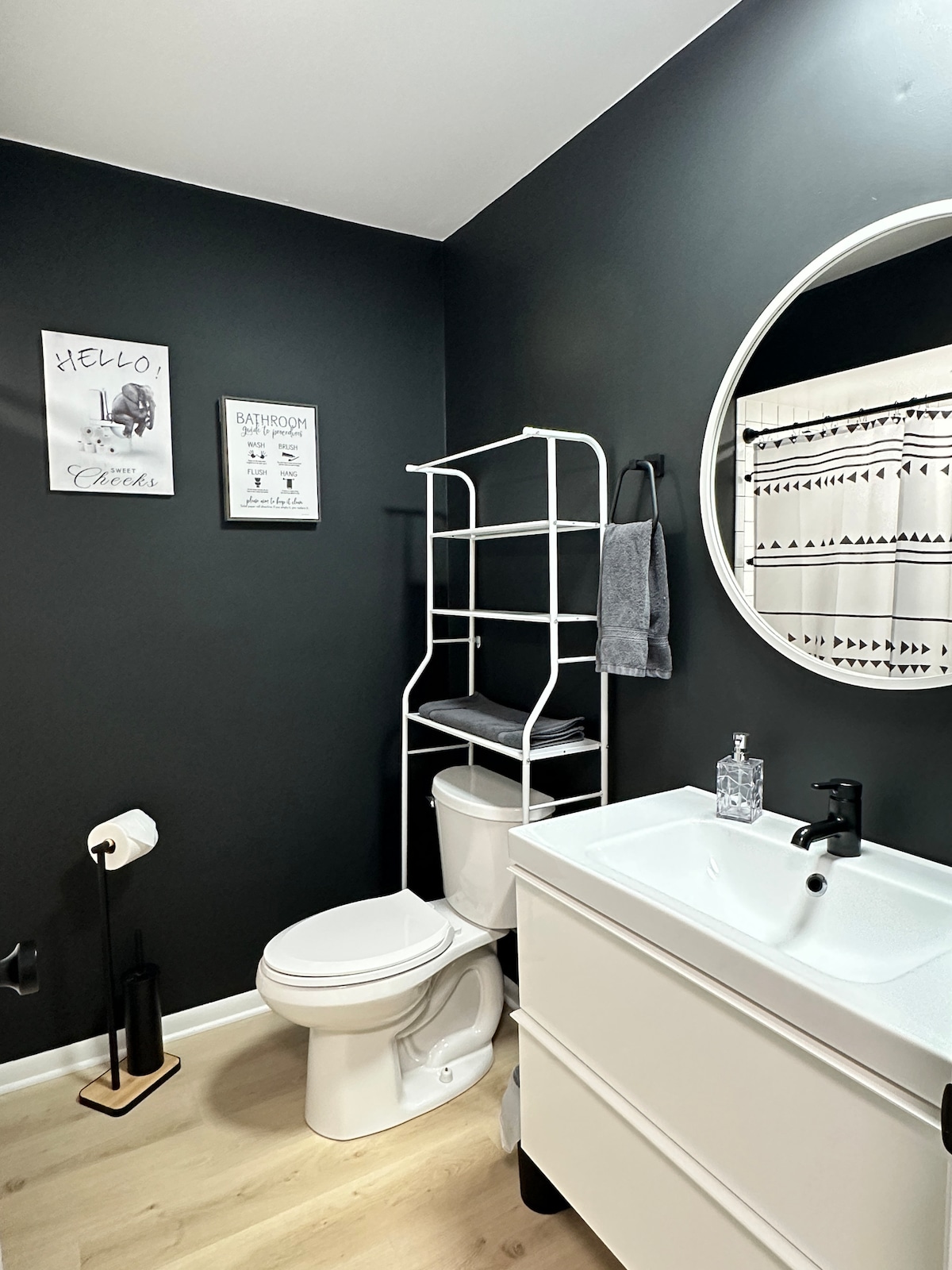 A modern bathroom is presented with dark walls and wooden flooring. A circular mirror reflects the space, while a white cabinet holds a sink. A toilet is positioned beside a ladder-style shelf, and coordinating shower curtains add a subtle pattern.